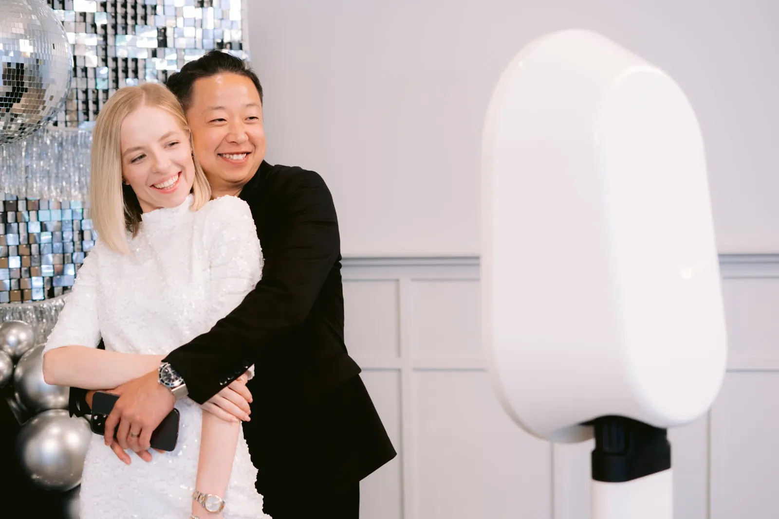 Newlywed couple posing together at a white open-air wedding photo booth with disco ball decor