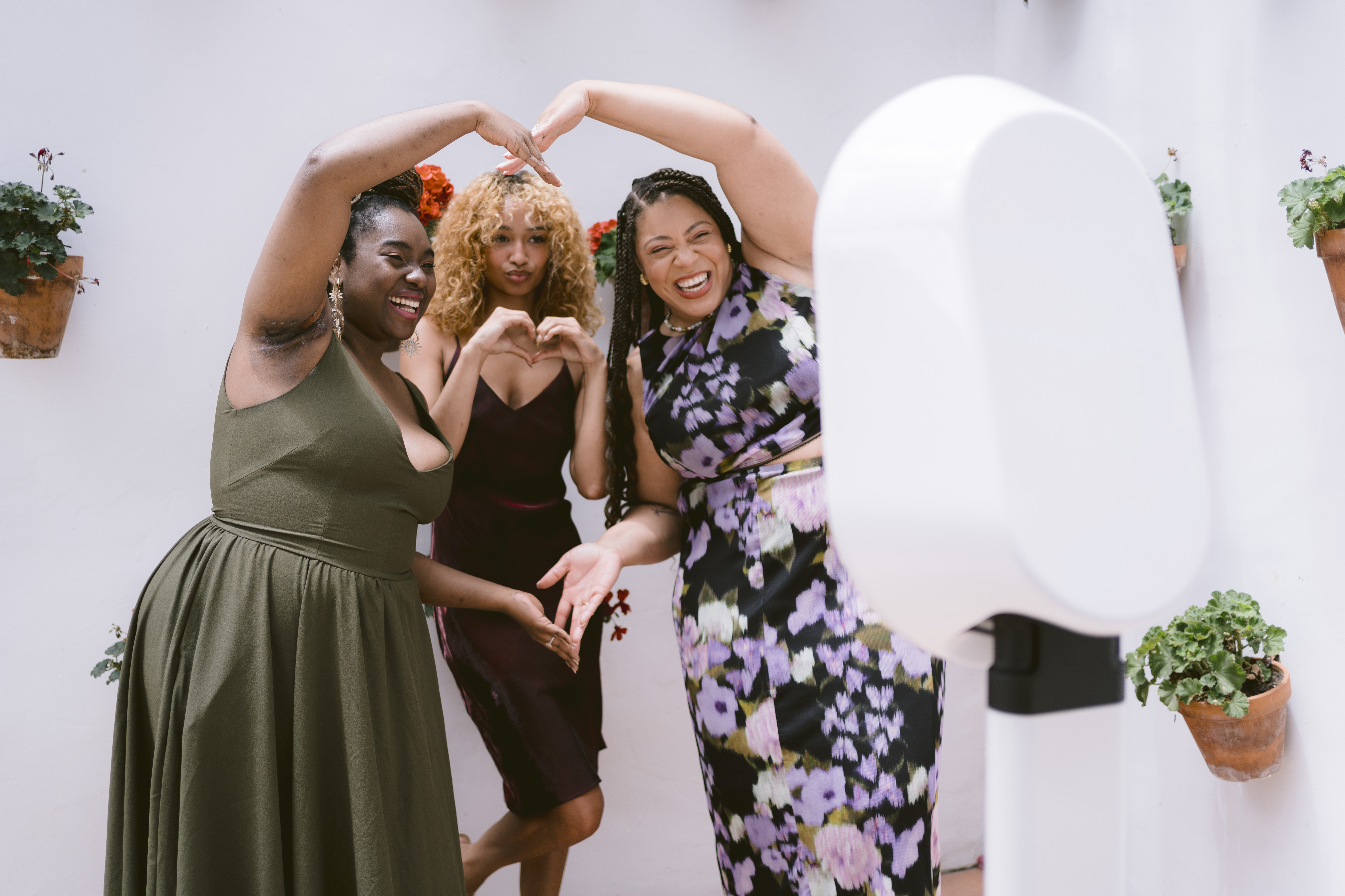 Three women posing with heart shapes in front of a photo booth, surrounded by potted plants on a white wall.