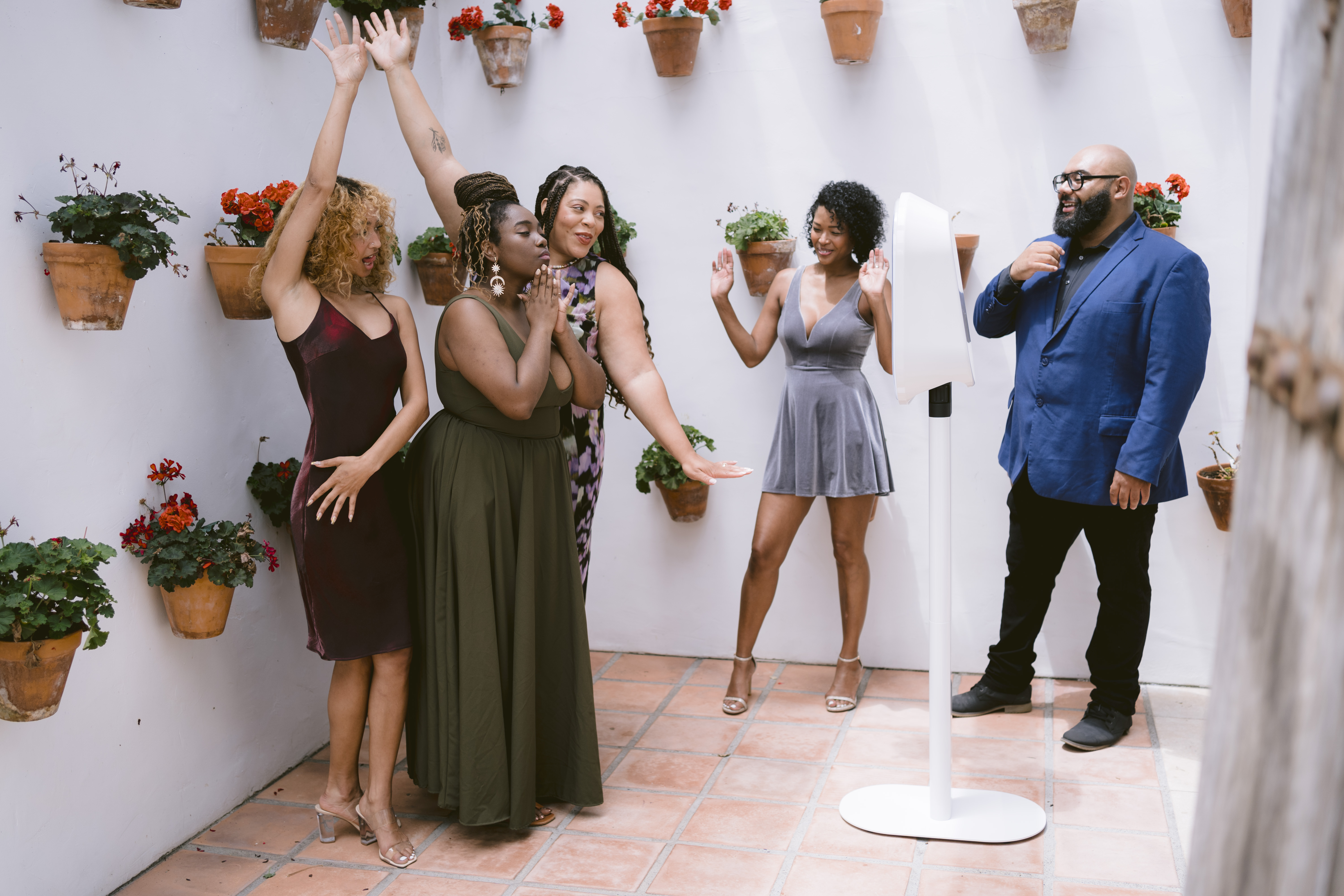 Group of diverse friends laughing and posing playfully with a modern white photo booth against a white wall with potted plants at an outdoor event.