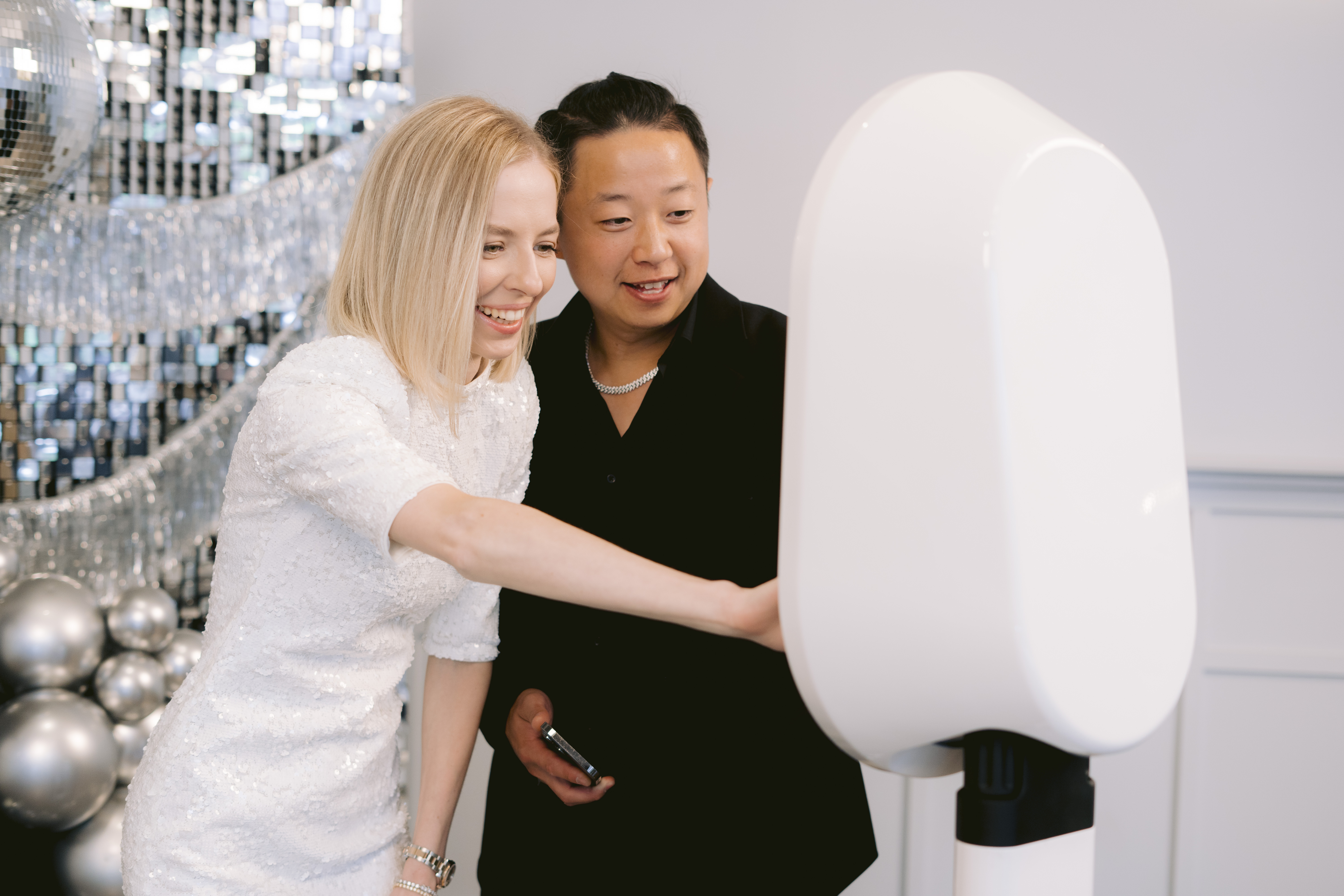 Smiling guests interacting with a modern white photo booth against a shimmering silver backdrop and balloons.